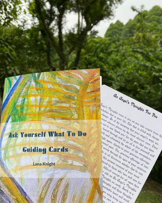 Ask Yourself What To Do Guiding Cards by Lana Knight with a colourful cover on the left. Behind it on the right is the card 'An Angel's Thoughts For you' fanned out. The card reads as ' An Angel's Thoughts come to you as you take respite' and continues. In the background there are green leaved trees shining under sun light. A painting of a multi coloured path way with foliage is used as the cover photo of the card deck.